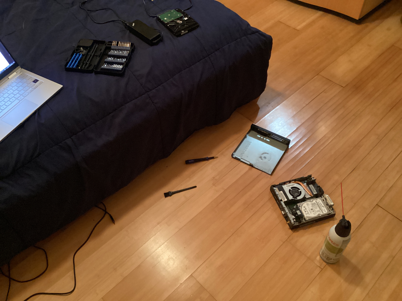 a picture of a machine slighly taken apart, with an air can and cable next to it. there is also a bed with a toolkit, a hard drive with a cable next to it, and a partially covered, turned on laptop.